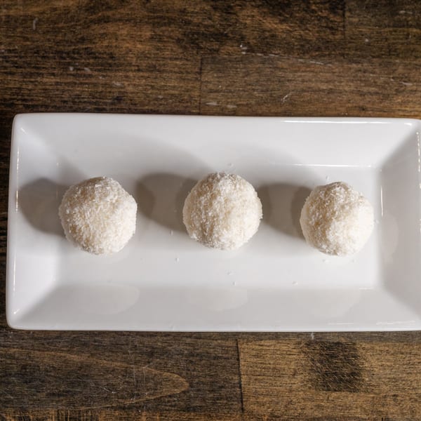 Three white coconut ladoos, Indian sweets, on a rectangular white plate on a wooden table.