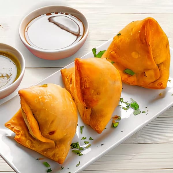 Three golden-brown Indian samosas garnished with herbs, served on a white plate with two dipping sauces.