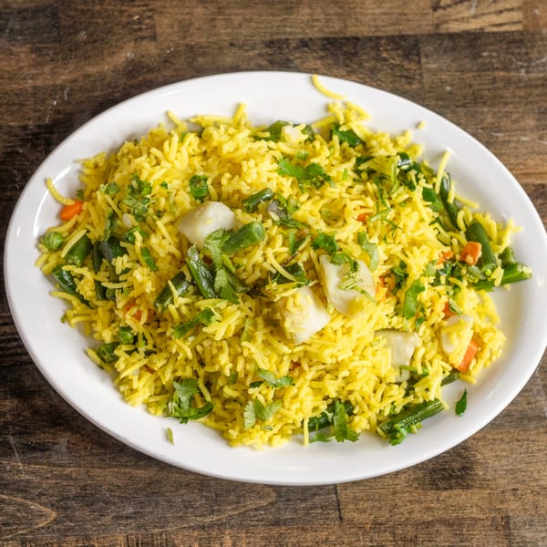 A white plate filled with bright yellow Indian rice, mixed with green beans, peas, carrots, and cilantro.