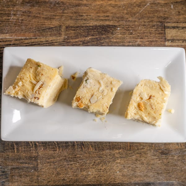 Three pieces of light yellow Indian Soan Papdi dessert with nuts on a white rectangular plate.