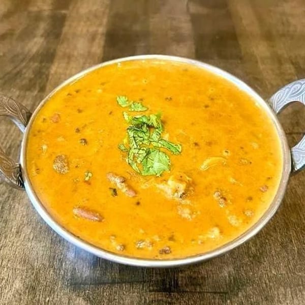 A metal bowl of orange Indian curry, garnished with fresh green herbs, on a wooden table.