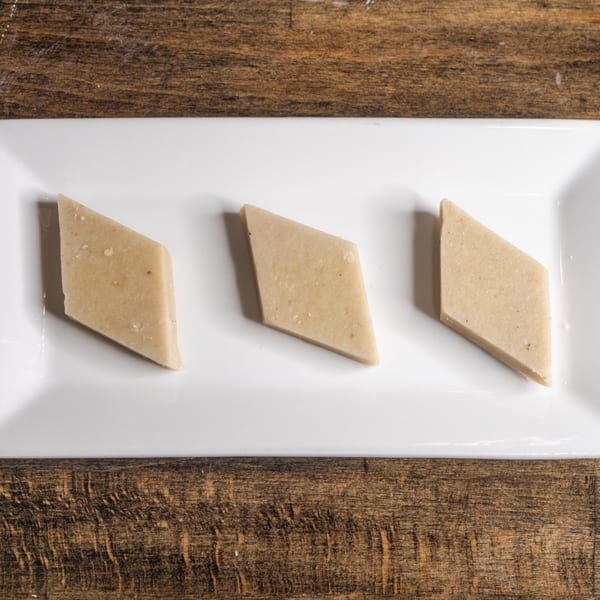 Three diamond-shaped pieces of light brown Indian barfi dessert on a white rectangular plate.